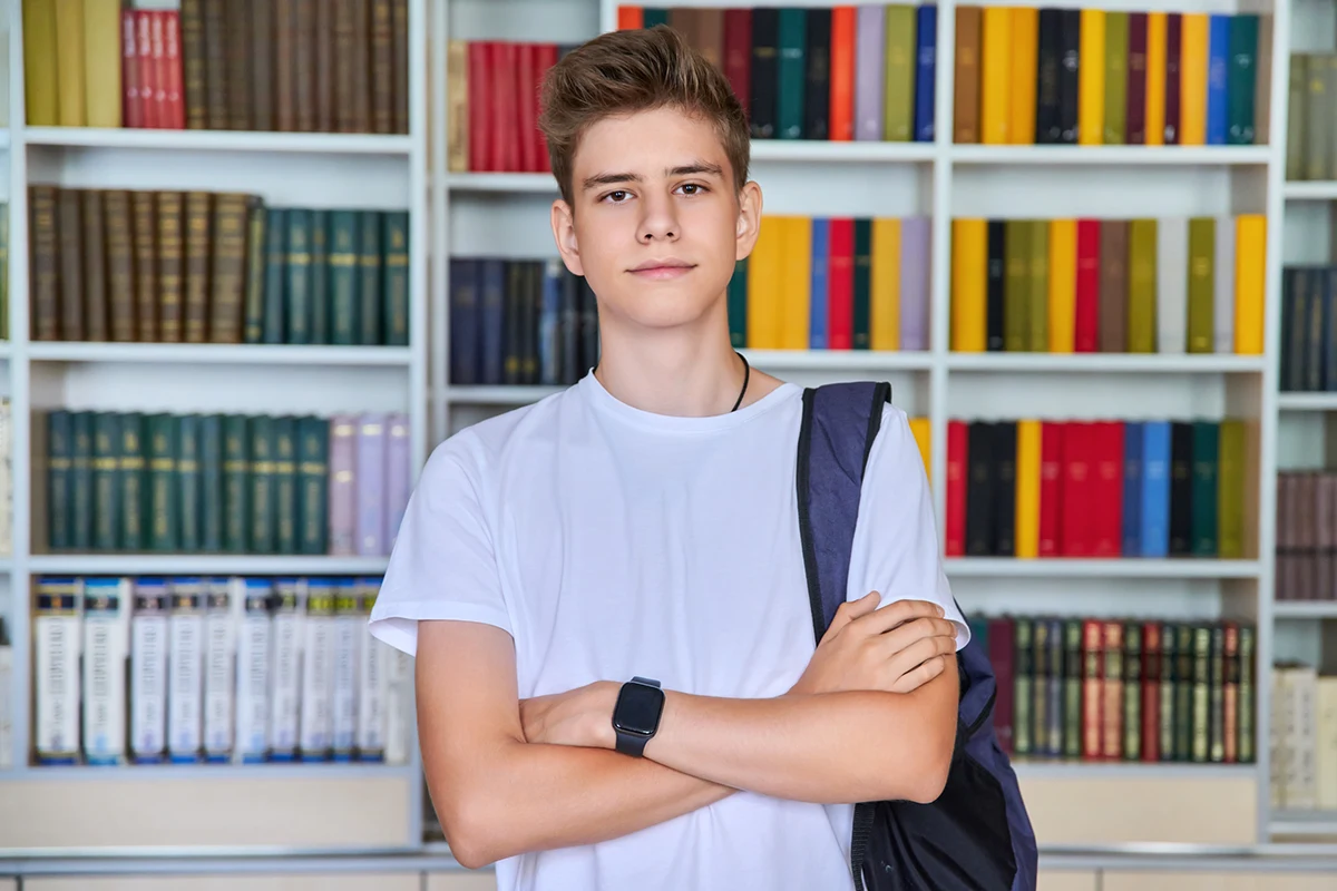 Best age for gynecomastia: Teenage boy standing with arms crossed in front of a bookshelf background